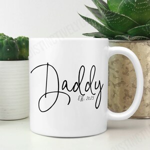May include: A white ceramic mug featuring the black script text Daddy and the smaller text Est. 2025 printed on the front. The mug has a standard cylindrical shape with a curved handle on the right side, presented in a clean and simple design.