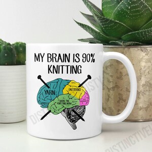 May include: White ceramic mug with the text "MY BRAIN IS 90% KNITTING" and a brain diagram showing knitting-related sections. The diagram includes "YARN," "PATTERNS," and "WHAT I'M KNITTING NOW." Two knitting needles cross the brain illustration.