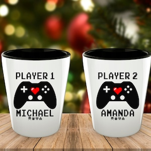 May include: Two white shot glasses with black rims. The glasses have a black and white pixelated design of a video game controller with a red heart. The text "Player 1" and "Michael" are on one glass, and the text "Player 2" and "Amanda" are on the other glass.