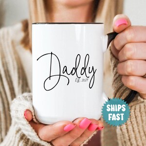 May include: A white ceramic mug with a black handle and interior. The front of the mug features the word Daddy in a black cursive script font, with the text Est. 2027 printed in a smaller, simple black font directly underneath the word.