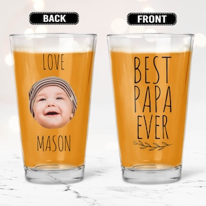 May include: A clear pint glass filled with a golden liquid. The front of the glass reads "BEST PAPA EVER" in black lettering with a small leaf design. The back of the glass has a baby's face and the name "MASON" below the image.