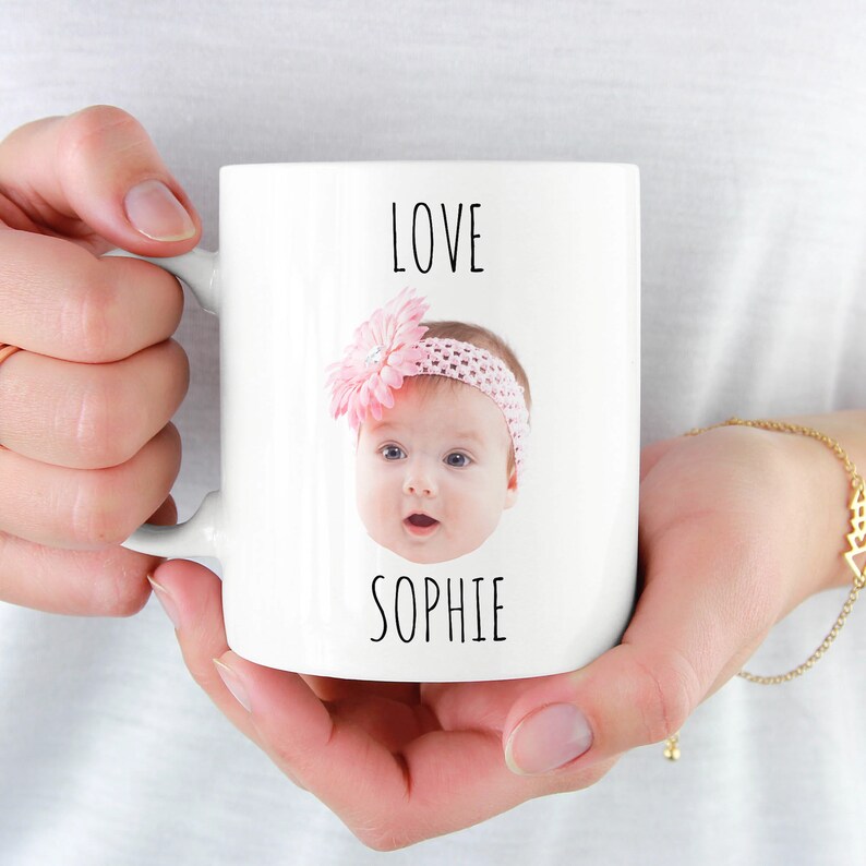 May include: White ceramic mug with a photo of a baby girl wearing a pink headband with a flower. The mug has the text "LOVE SOPHIE" printed on it.