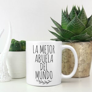 May include: White ceramic mug with the text "LA MEJOR ABUELA DEL MUNDO" in black, with a sprig of leaves at the bottom. The mug is on a white surface, with plants in the background.