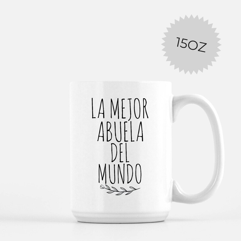 May include: White ceramic mug with the text "LA MEJOR ABUELA DEL MUNDO" in black. The mug has a capacity of 15 oz. A sprig of leaves is printed below the text. The mug has a large handle.