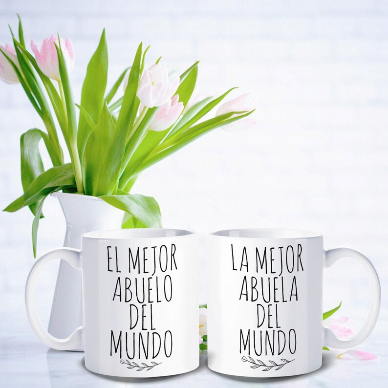 May include: Two white ceramic mugs with black text that reads "EL MEJOR ABUELO DEL MUNDO" and "LA MEJOR ABUELA DEL MUNDO". The mugs are in front of a white vase with pink and white tulips.