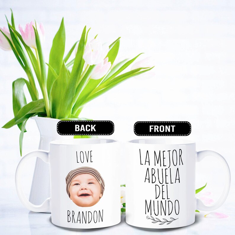 May include: White ceramic mug with a photo of a baby and the name "Brandon" on one side, and the text "LA MEJOR ABUELA DEL MUNDO" on the other. The mug is set against a backdrop of white and a vase of pink and white tulips.