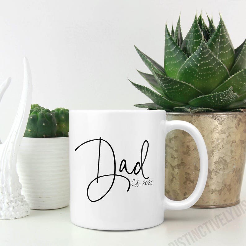 May include: A white ceramic mug featuring the black cursive text "Dad" followed by "Est. 2026" in a smaller, simple font. The mug has a standard cylindrical shape with a curved handle on the right side, presented against a plain white background.