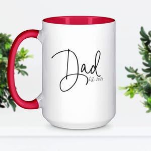 May include: A white ceramic mug with a bright red interior and a matching red handle. The front of the mug features the word Dad in a black cursive script font, with the text Est. 2026 printed in a smaller, simple black font underneath it.