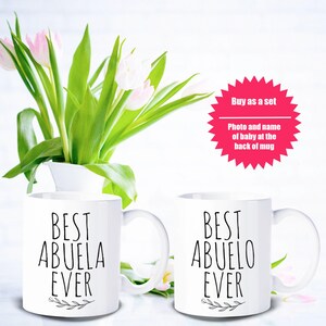 May include: Two white ceramic mugs with the text "BEST ABUELA EVER" and "BEST ABUELO EVER" in black script. The mugs are displayed with a bouquet of pink and white tulips in a white vase. A pink starburst graphic reads "Buy as a set".