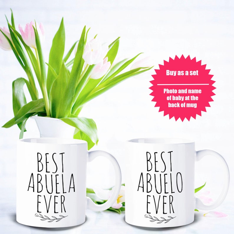 May include: Two white ceramic mugs with the text "BEST ABUELA EVER" and "BEST ABUELO EVER" in black script. The mugs are displayed with a bouquet of pink and white tulips in a white vase. A pink starburst graphic reads "Buy as a set".