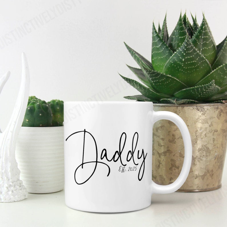 May include: A white ceramic mug featuring the black script text Daddy and the smaller text Est. 2025 printed on the front. The mug has a standard cylindrical shape with a curved handle on the right side, presented in a clean and simple design.