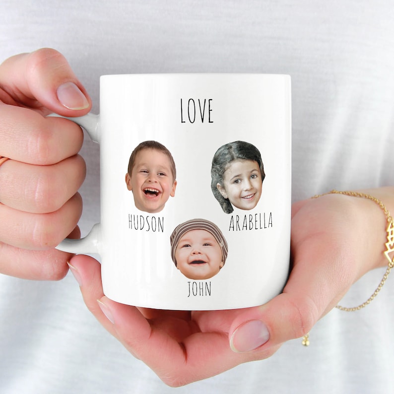 May include: White ceramic mug featuring three smiling faces with the names Hudson, Arabella, and John below each. The word "LOVE" is printed above the faces. The mug is held by a person.