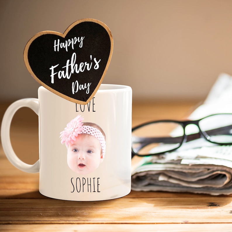 May include: White ceramic mug with a photo of a baby girl wearing a pink headband and a pink flower. The mug has the text "LOVE" and "SOPHIE" printed on it. A black heart-shaped sign with the text "Happy Father's Day" is on top of the mug.