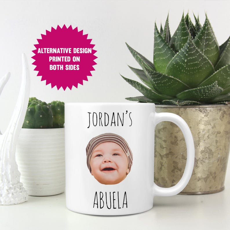 May include: White ceramic mug featuring a smiling baby's face, the name "Jordan's" above the image, and the word "ABUELA" below. The mug has a curved handle and the text "ALTERNATIVE DESIGN PRINTED ON BOTH SIDES" on a pink starburst.