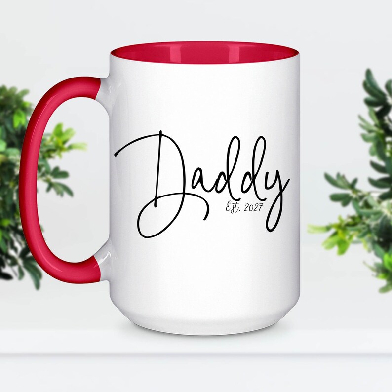 May include: A white ceramic mug with a bright red handle and interior rim. The front of the mug features the word Daddy in a black cursive script font, with the text Est. 2027 printed in a smaller, simple black font underneath the word.