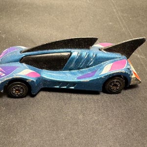 May include: A blue toy car with black fins and black wheels. The car has purple, pink, and white accents. The toy car is a die-cast model.