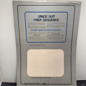 May include: A gray instruction label with a blue border, titled "SPACE SUIT PREP SEQUENCE." The label includes text about preparing a spacesuit, with numbered steps and warnings. The bottom of the label reads "INSTRUCTION LABELS."
