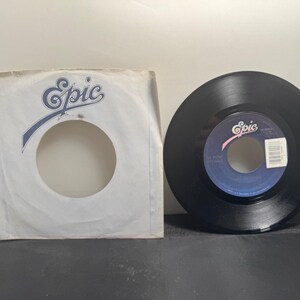 May include: A black vinyl record with a blue and white label, next to a white paper sleeve. The label and sleeve both feature the word "Epic" in a stylized font. The record is a 45 RPM single.