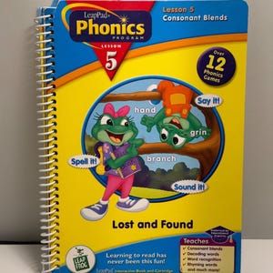 May include: A spiral-bound LeapPad Phonics Program Lesson 5 book titled Lost and Found. The yellow cover features cartoon frog characters, the text Lesson 5 Consonant Blends, and a box listing educational goals like decoding and word recognition.