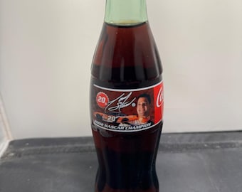 May include: A Coca-Cola bottle with a red cap and a green neck. The bottle has a label featuring a NASCAR driver and the text "2002 NASCAR CHAMPION". The bottle is filled with a dark liquid.