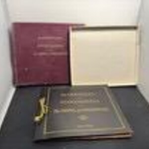 May include: A collection of vintage books with burgundy and brown covers. One book is open, revealing cream-colored pages. The open book has a gold clasp and the title is visible. The books are in a box.