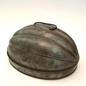 May include: A vintage, oval-shaped, metal cake mold with a fluted design and a wire handle. The mold is made of tin and has a dark gray patina.
