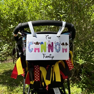 Op de afbeelding: Een wit bord met zwarte tekst waarop "The Cannon Family" staat, met kleurrijke letters versierd met cartoonfiguren. Het bord is bevestigd aan een zwarte kinderwagen met rode, gele en zwarte linten.