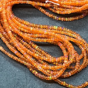 May include: A strand of orange opal beads, with iridescent highlights, is shown in a close-up image. The beads are small and round, and they are strung together on a clear thread.