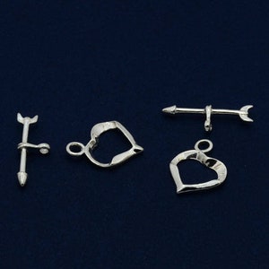 Op de afbeelding: Drie zilverkleurige metalen sluitingen voor het maken van sieraden. Eén sluiting is hartvormig, de andere twee zijn pijlvormig.