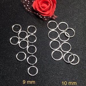 Op de afbeelding: Twee sets zilveren springringen, een set van 9 mm ringen en een set van 10 mm ringen. De ringen zijn in een cirkelvormige opstelling op een zwarte achtergrond.