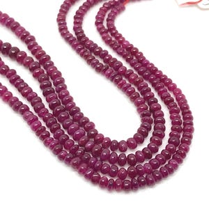 May include: Four strands of ruby gemstone beads are shown against a white backdrop. The beads are a deep, rich red-purple colour, and each bead is round. The strands are strung together with a red string.