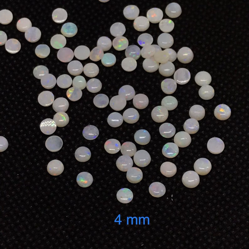 Pu&ograve; includere: Una vista ravvicinata di un mucchio di cabochon di opale bianco, ognuno con un diametro di 4 mm. Le pietre hanno un sottile bagliore iridescente.