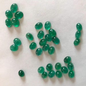 May include: A collection of oval-shaped, green gemstones. The gemstones are arranged in a variety of patterns on a white surface.