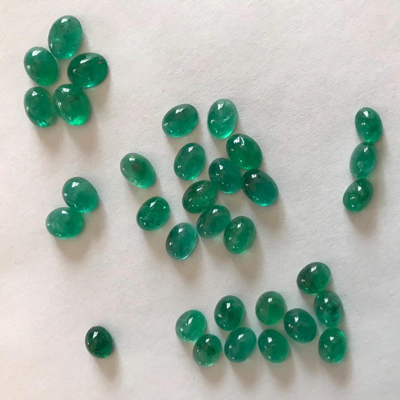 May include: A collection of oval-shaped, green gemstones. The gemstones are arranged in a variety of patterns on a white surface.