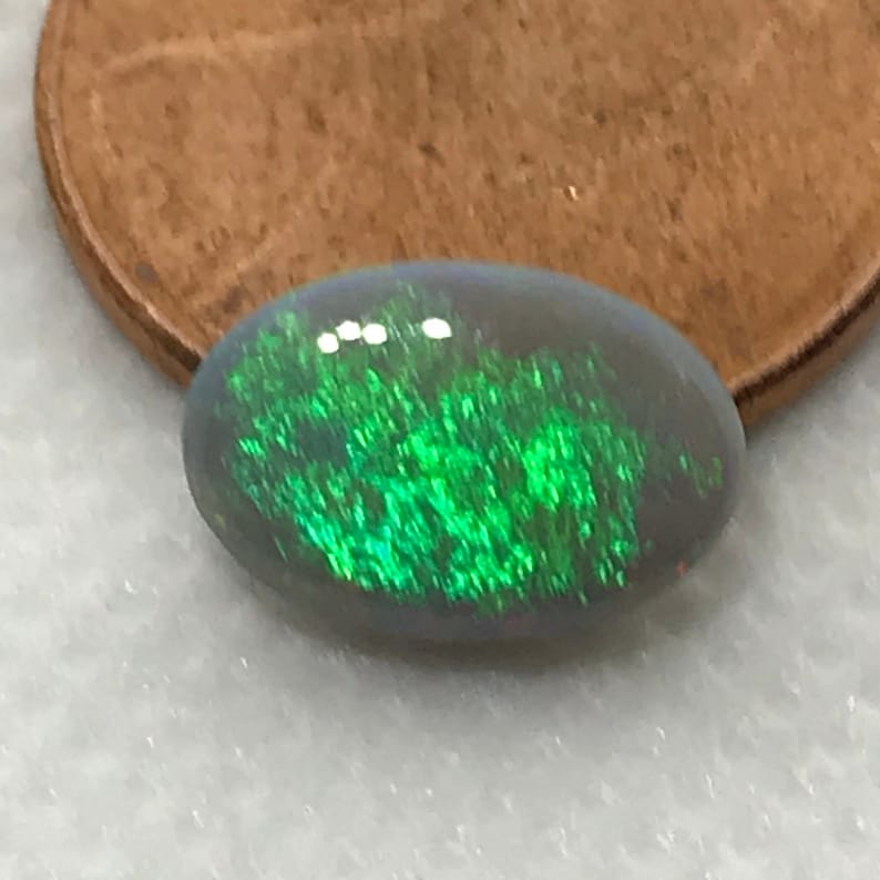May include: A green and grey oval opal gemstone with a bright green iridescent sheen. The opal is about the size of a UK penny.