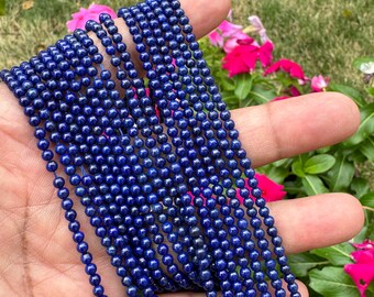 Könnte beinhalten: Eine Strähne tiefblauer Lapislazuli-Perlen, etwa 4 mm im Durchmesser, auf einem dünnen Faden aufgereiht. Die Perlen sind rund und glatt, mit einer leichten Variation in Farbe und Muster. Die Strähne wird in einer Hand gehalten, der Hintergrund ist verschwommen.