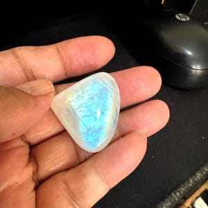 May include: A polished, iridescent moonstone gemstone, displaying a play of blue and yellow light. The stone is held in a hand, showcasing its smooth, rounded shape and natural beauty. The gemstone's surface reflects light, creating a shimmering effect.