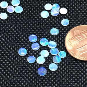 Könnte beinhalten: Eine Sammlung kleiner, runder, irisierender Opal-Cabochons, die auf einer schwarzen Oberfläche verstreut sind. Die Opale sind in verschiedenen Blautönen und Weißtönen gehalten, einige zeigen grüne und rote Schimmer. Ein Cent-Stück ist zur Größenbestimmung enthalten.