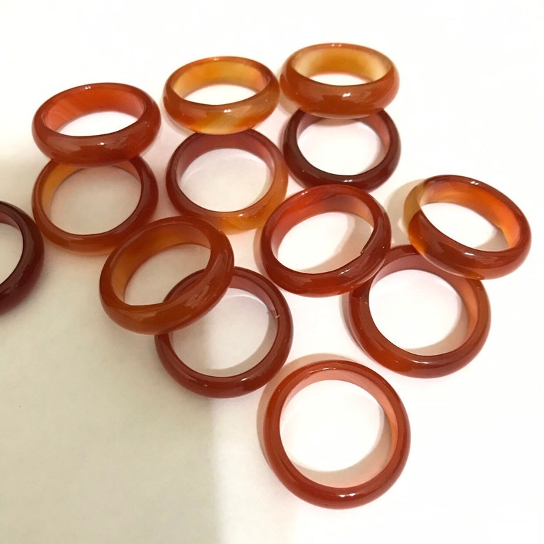 May include: A group of 12 carnelian stone rings. The rings are a deep orange color with a slight reddish hue. They are all smooth and polished.