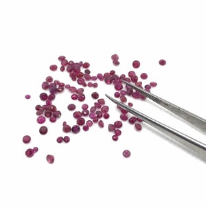 May include: A scattering of small, round, red gemstones on a white surface. A pair of tweezers is lying on the surface, near the gemstones.