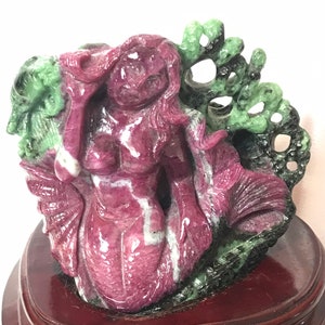 May include: A pink and green carved jade mermaid figurine on a wooden base. The mermaid is sitting on a rock with her tail curled around her legs. The rock is carved with a floral design.