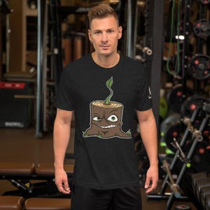 Peut inclure: T-shirt gris foncé avec un dessin de souche d'arbre de dessin animé. La souche a une pousse, une bouche souriante et un seul œil visible. Le dessin est souligné en blanc et vert. Le t-shirt est à manches courtes et en matière douce.
