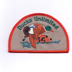 May include: A tan and orange embroidered patch with a red border. The patch features a duck with a green head and a brown body. The text "Ducks Unlimited" is embroidered in green on the patch.