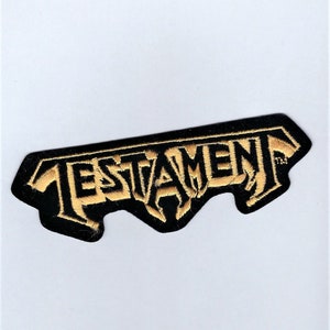 May include: Black and gold embroidered patch with the word "TESTAMENT" in a stylized font.