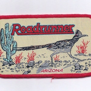 May include: An embroidered patch in red, white and blue featuring the words "Roadrunner" and "Arizona". The patch depicts a cartoon roadrunner with a long, thin beak and a long tail. The roadrunner is standing on a desert landscape with a cactus and bushes.