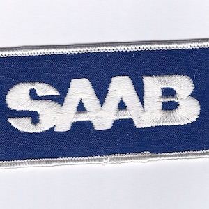 May include: A white embroidered patch with the word "SAAB" in white lettering on a dark blue background. The patch is surrounded by a white border.