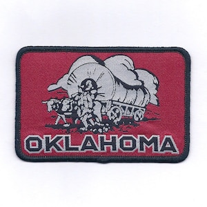 May include: A red embroidered patch with a black outline. The patch features a black and white illustration of a covered wagon being pulled by two oxen. The text "OKLAHOMA" is printed in silver below the wagon.