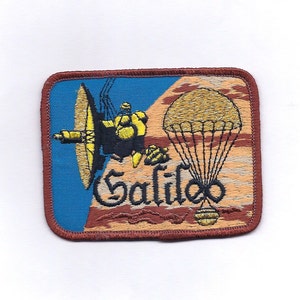 May include: Embroidered patch featuring a spacecraft and a parachute design with the name "Galileo". The patch has a blue background with a brown border and a yellow and brown spacecraft.