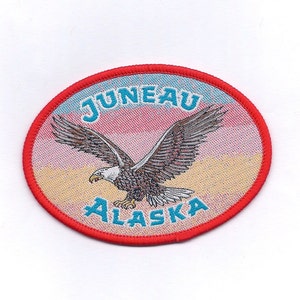 May include: An oval-shaped embroidered patch with a red border. The patch features a bald eagle in flight with the words "JUNEAU ALASKA" in blue lettering.