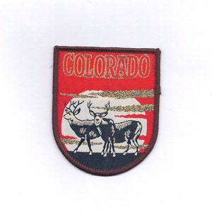 May include: A brown and red embroidered patch with the word "COLORADO" in gold lettering. The patch features two black silhouette deer standing in front of a white moon with a gold glitter background.